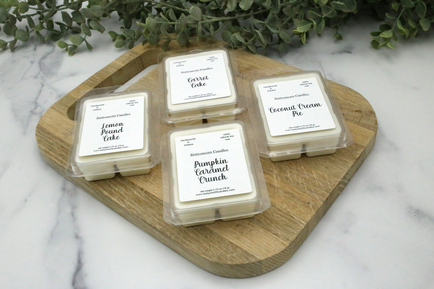 Bakery-scented wax melts from Sixtyone20 Candles in Pumpkin Caramel Crunch, Coconut Cream Pie, Carrot Cake, and Lemon Pound Cake, displayed on a wooden cutting board with greenery in the background.