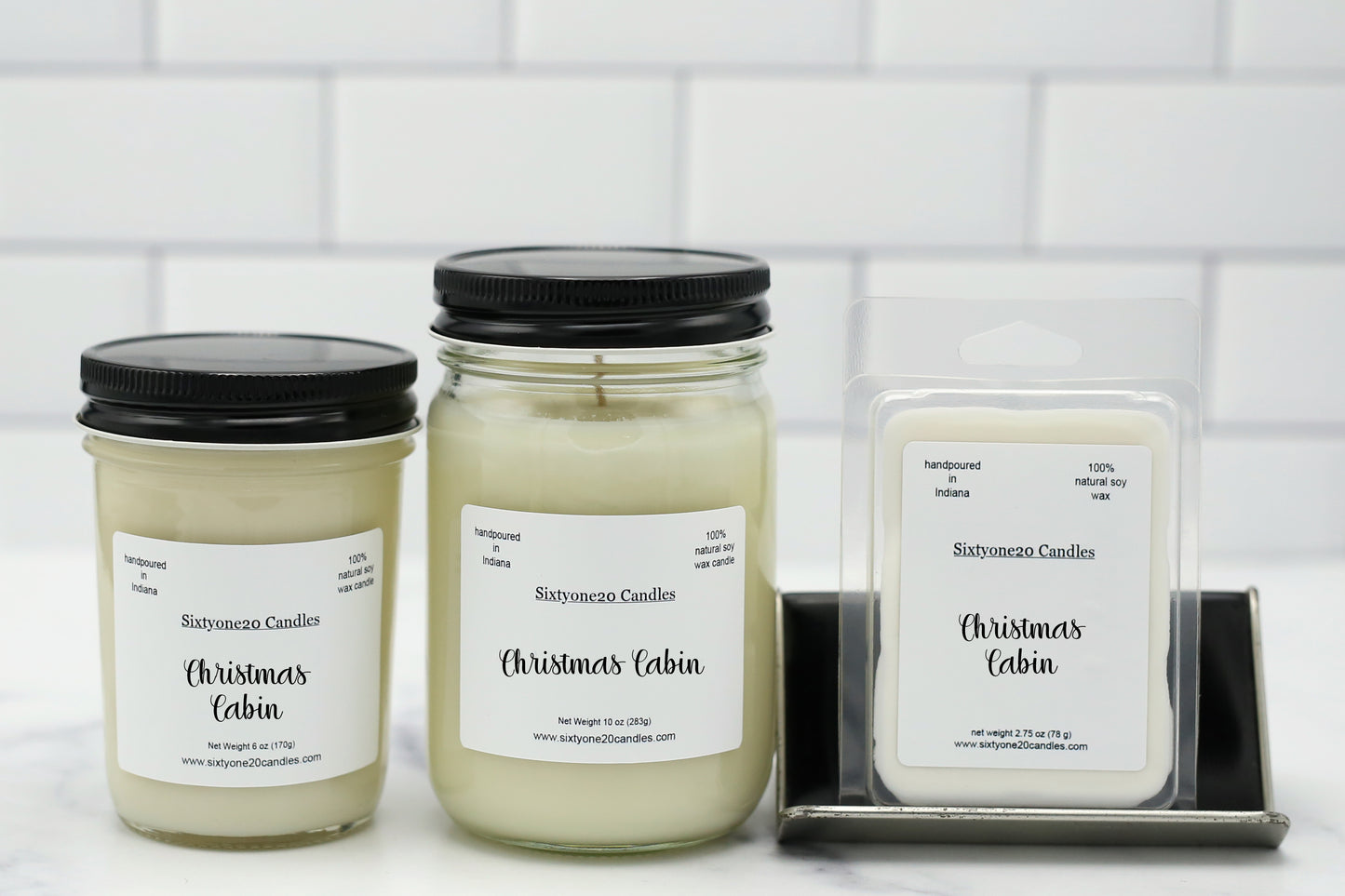 Three Sixtyone20 Candles 'Christmas Cabin' scented products displayed on a white surface against a white subway tile background: two glass jar candles with black lids and one wax melt package in a clear plastic clamshell on a small metal stand.