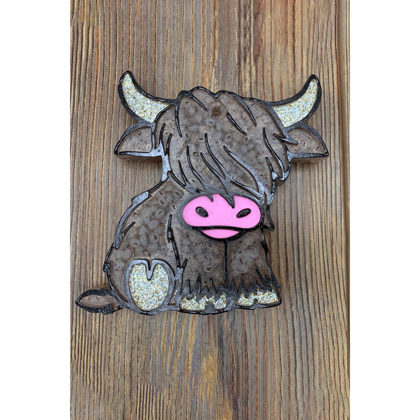 Sitting Highland cow-shaped freshie with glittery horns and hooves, long brown fur, and a pink snout, placed on a wood background.