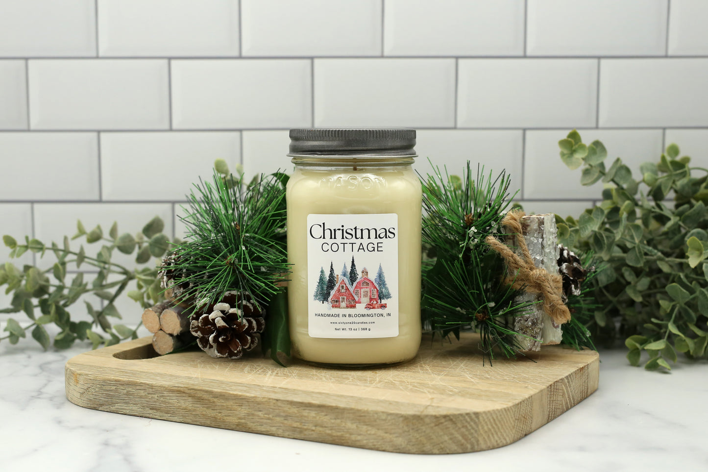 Christmas Cottage Winter Candle Club exclusive in 16 oz mason jar with silver lid, styled with pinecones and greenery.