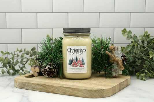 Christmas Cottage Winter Candle Club exclusive in 16 oz mason jar with silver lid, styled with pinecones and greenery.