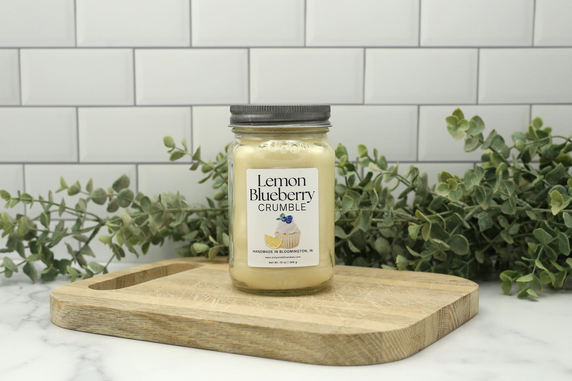 Spring 2026 Candle Club Lemon Blueberry Crumble candle in a 16 oz mason jar with silver lid on a wooden board with greenery background