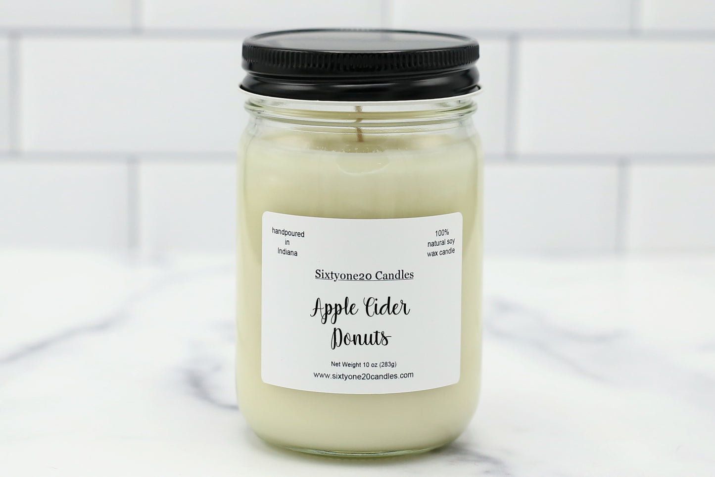A glass jar candle with a black lid and a white label reading 'Sixtyone20 Candles Apple Cider Donuts,' set on a light marble surface with a white subway tile background.