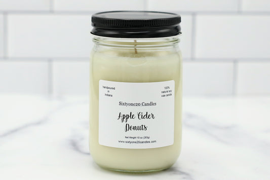 A glass jar candle with a black lid and a white label reading 'Sixtyone20 Candles Apple Cider Donuts,' set on a light marble surface with a white subway tile background.