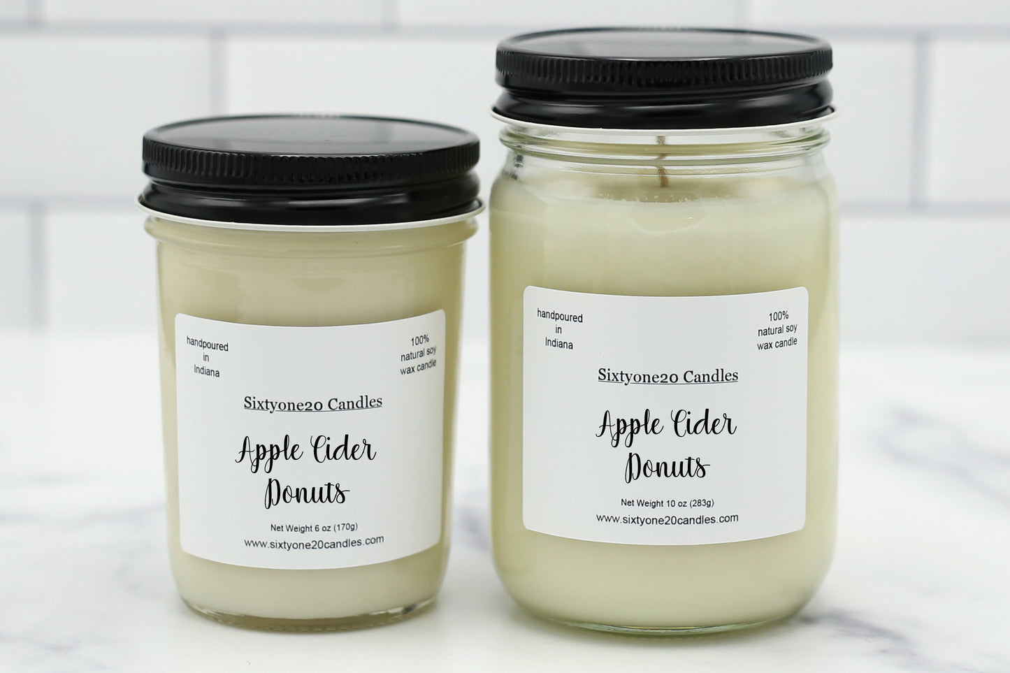 Two glass jar candles with black lids and white labels reading 'Sixtyoneo Candles Apple Cider Donuts' on a light marble surface with a white subway tile background.