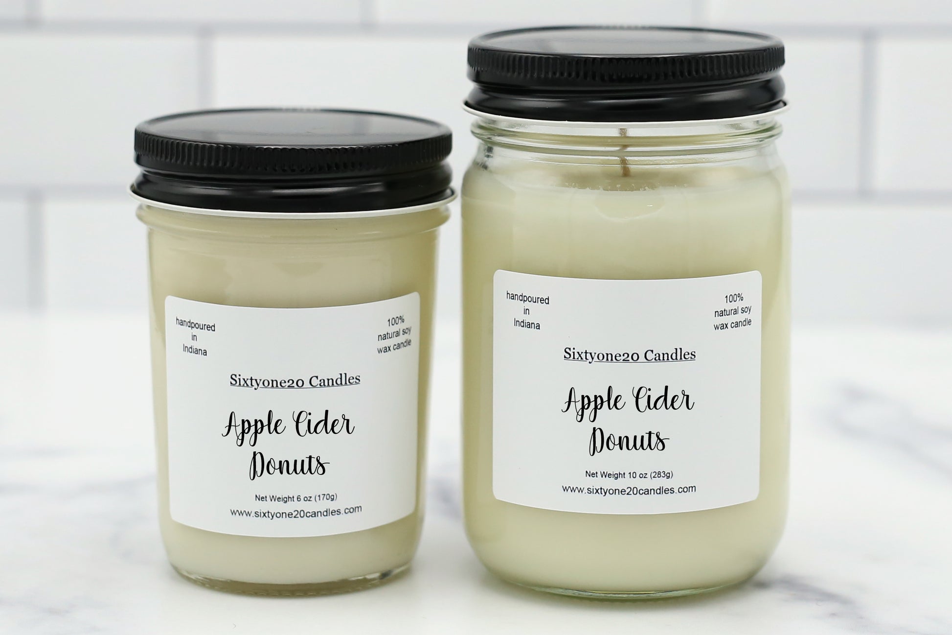 Two glass jar candles with black lids and white labels reading 'Sixtyoneo Candles Apple Cider Donuts' on a light marble surface with a white subway tile background.
