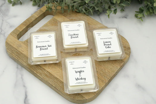 Wax melts from Sixtyone20 Candles in Banana Nut Bread, Lemon Pound Cake, Waffles & Whiskey, and Zucchini Bread, arranged on a wooden cutting board with a greenery backdrop.