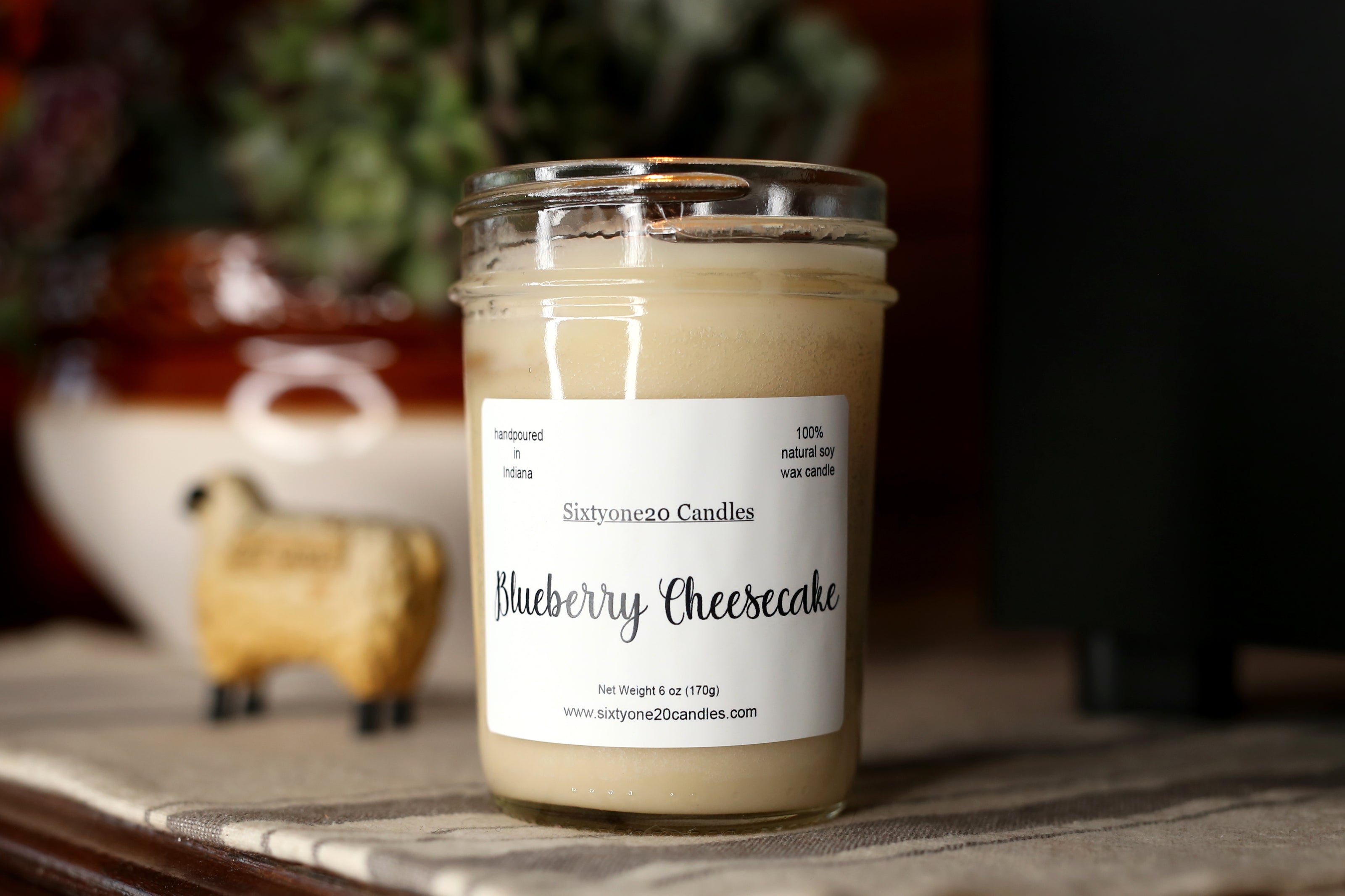 Candle labeled 'Blueberry Cheesecake' by Sixtyone20 Candles on a surface with a blurred background