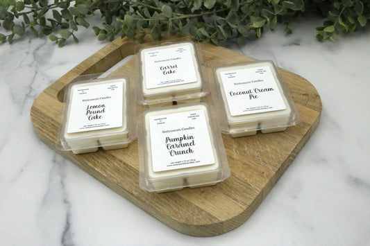 Bakery-scented wax melts from Sixtyone20 Candles in Pumpkin Caramel Crunch, Coconut Cream Pie, Carrot Cake, and Lemon Pound Cake, displayed on a wooden cutting board with greenery in the background.