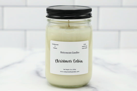 A glass jar candle with a black lid and a white label reading 'Sixtyone20 Candles Christmas Cabin,' set on a light marble surface with a white subway tile background.