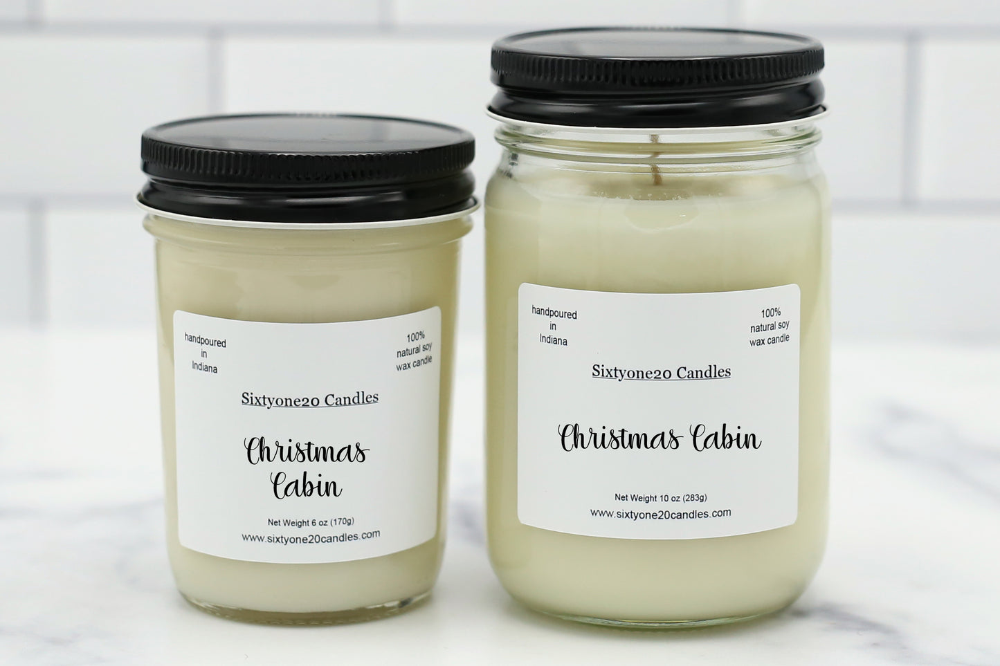 Two glass jar candles with black lids and white labels reading 'Sixtyoneo Candles Christmas Cabin' on a light marble surface with a white subway tile background.