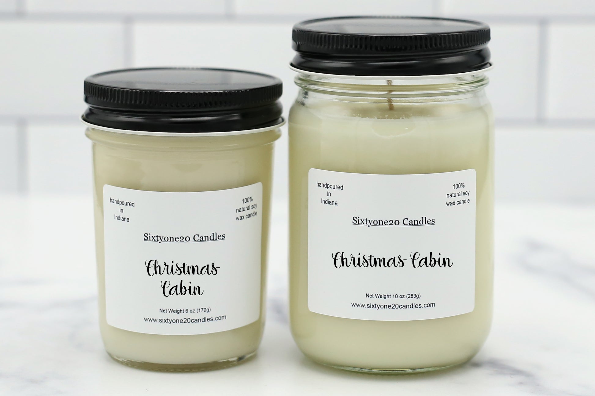 Two glass jar candles with black lids and white labels reading 'Sixtyoneo Candles Christmas Cabin' on a light marble surface with a white subway tile background.