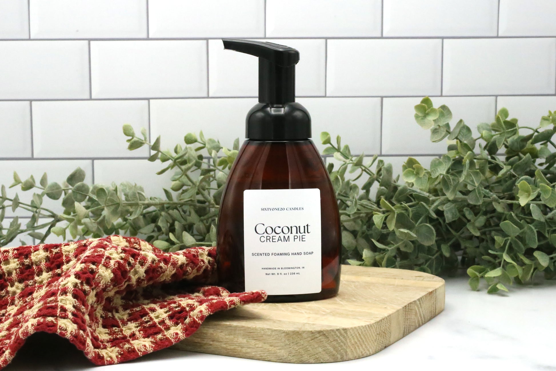 Coconut Cream Pie foaming hand soap by Sixtyone20 Candles displayed on a wooden board with red kitchen towel and greenery background