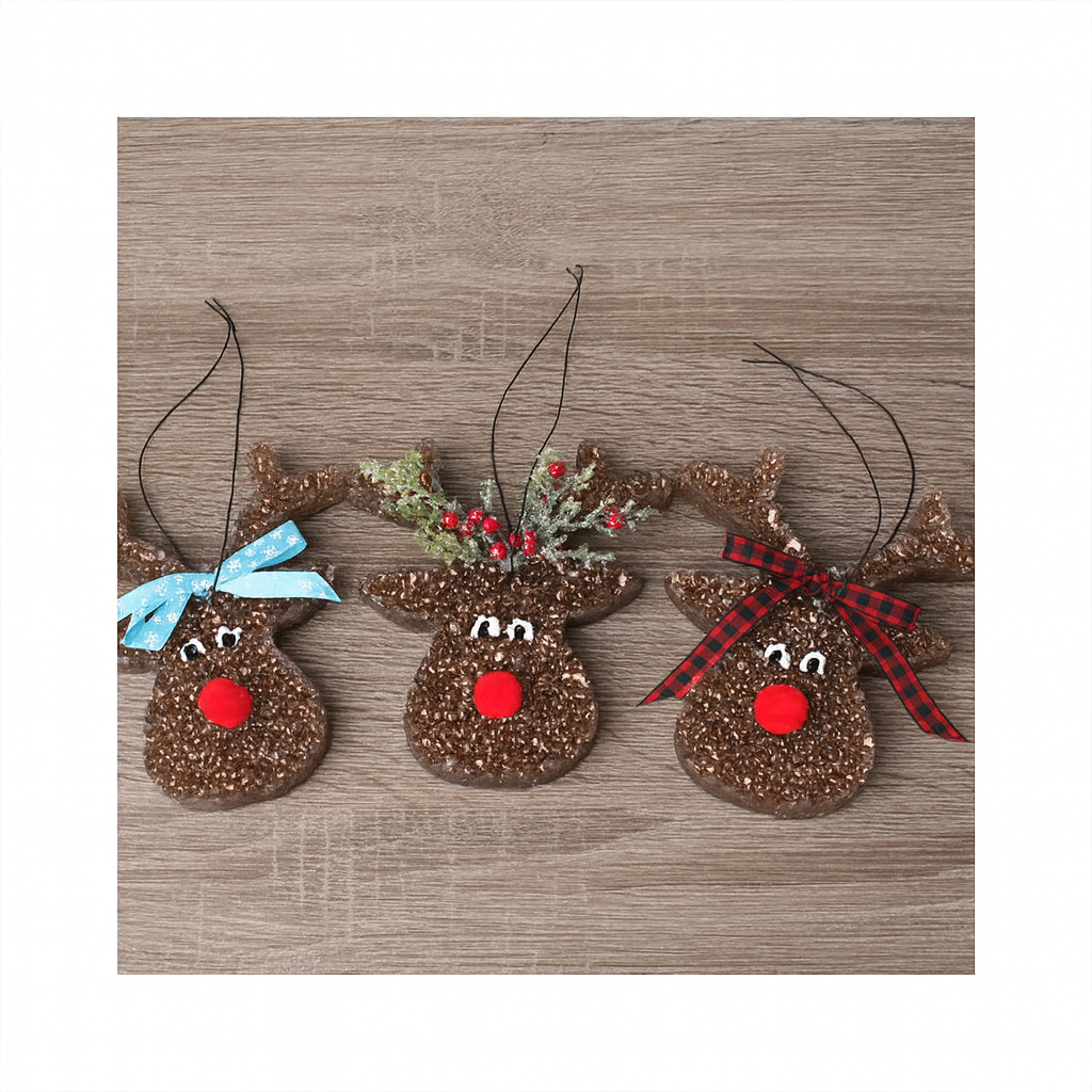 Three brown reindeer-shaped freshies with sparkly texture are arranged side by side on a wooden surface. Each has a red pom-pom nose and painted white eyes. The left reindeer has a light blue snowflake bow, the middle one has greenery with red berries, and the right one has a red and black plaid bow. White borders frame the image for a square format.