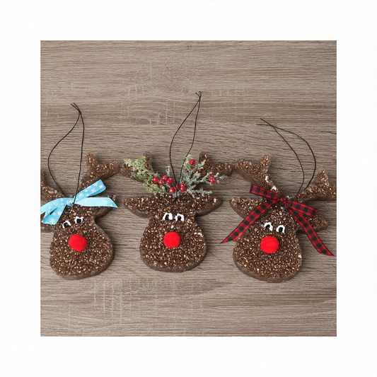 Three brown reindeer-shaped freshies with sparkly texture are arranged side by side on a wooden surface. Each has a red pom-pom nose and painted white eyes. The left reindeer has a light blue snowflake bow, the middle one has greenery with red berries, and the right one has a red and black plaid bow. White borders frame the image for a square format.